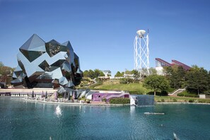 A modern building with a unique geometric facade near a body of water, a tall white tower, and a staircase leading up to a red-roofed structure.