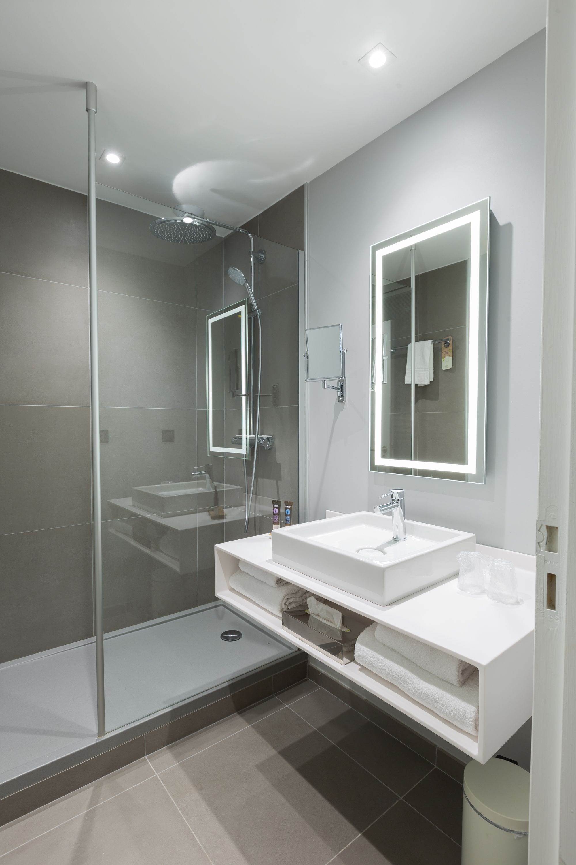 A modern bathroom with a glass-enclosed shower, a white sink, and a large mirror with built-in lighting.