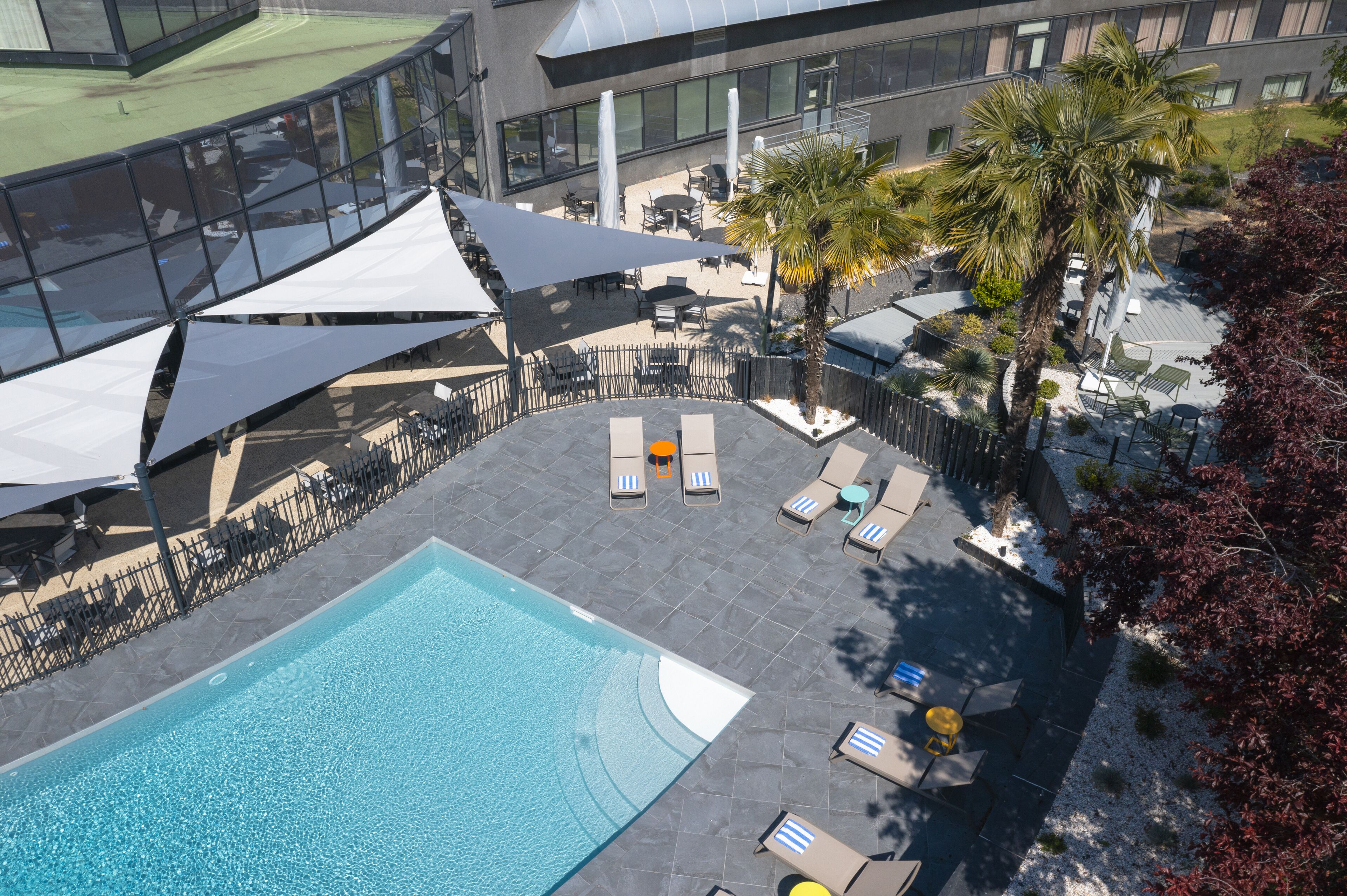 Un área de piscina en la azotea con sillones de descanso y sombrillas, rodeada por un edificio moderno con grandes ventanales.
