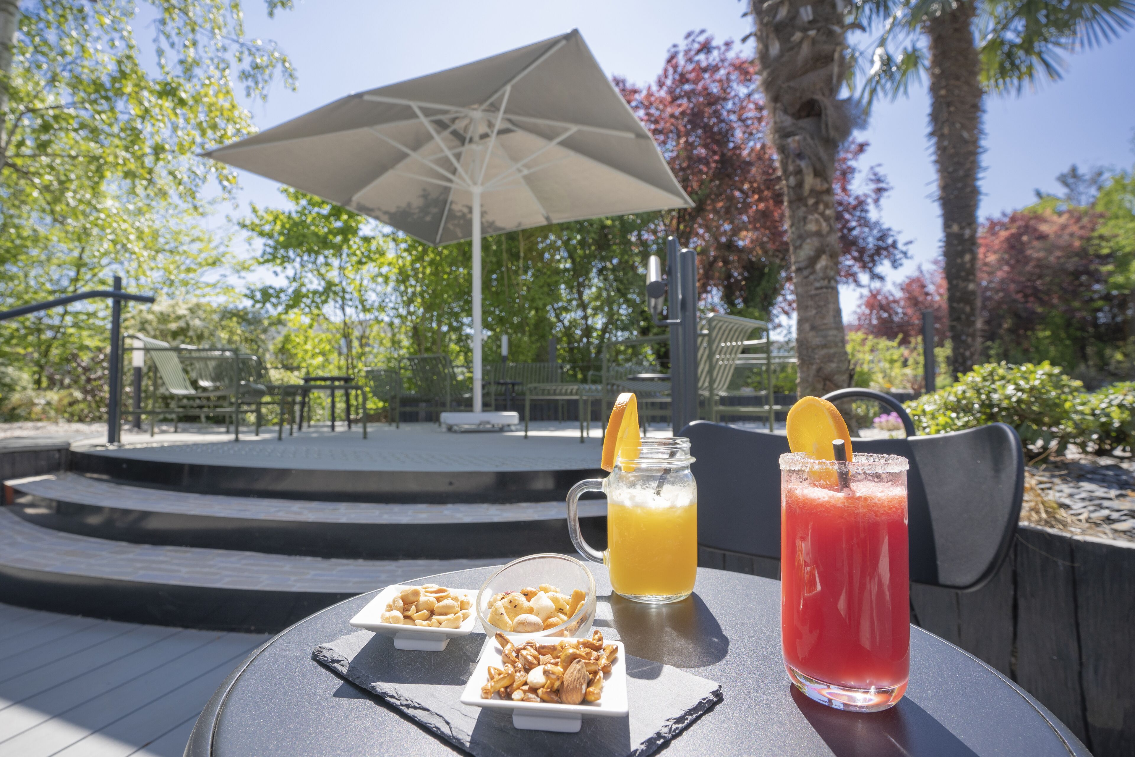 Ein Tisch mit Getränken, Nüssen und Obst, draußen mit Sonnenschirm und Stühlen im Hintergrund.