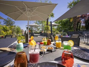 An outdoor dining area with tables, chairs, umbrellas, and various beverages. - Novotel Poitiers Site du Futuroscope (Chasseneuil-du-Poitou)