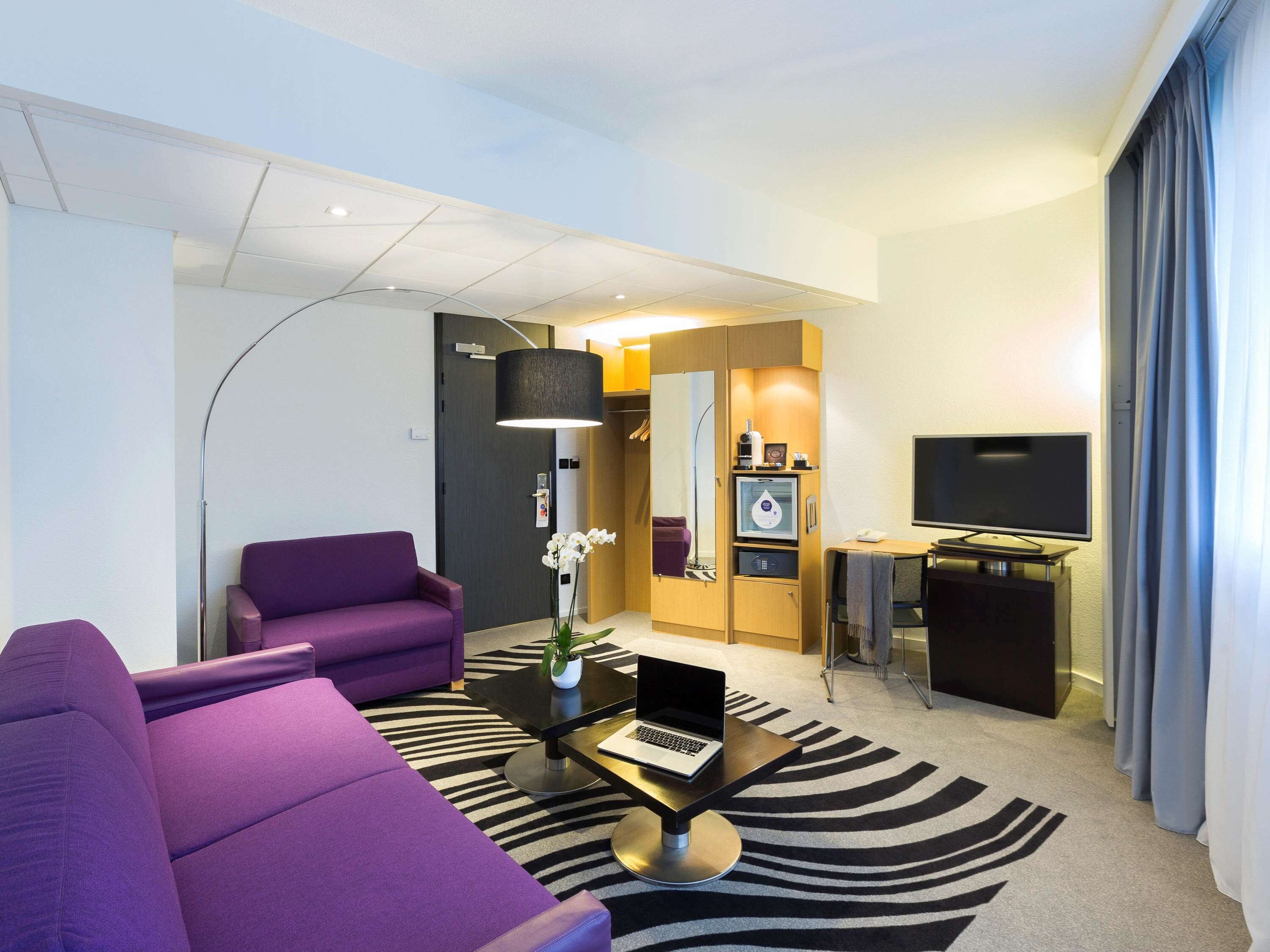 A modern hotel room with a purple sofa, a laptop on a table, a TV, and a minibar.
