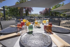 An outdoor dining area with tables set for two, featuring a variety of beverages and a fruit basket.