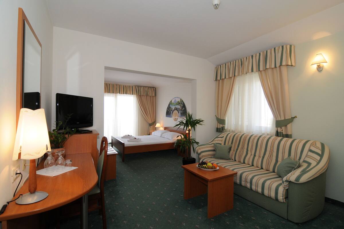 A hotel room with a bed, a sofa, a dining table, a TV, and a lamp.