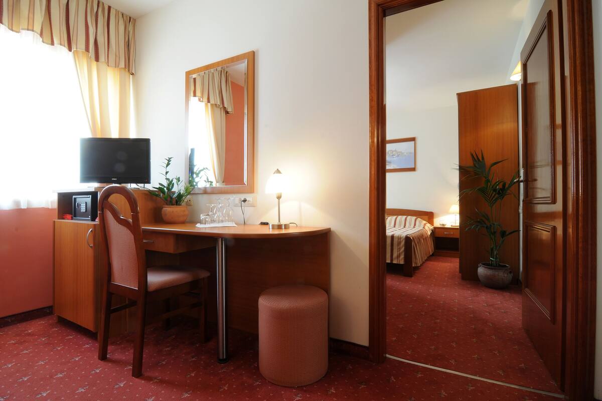 A hotel room with a wooden desk, a chair, a small stool, a television, a lamp, a mirror, a bed with a red carpet, and a potted plant.