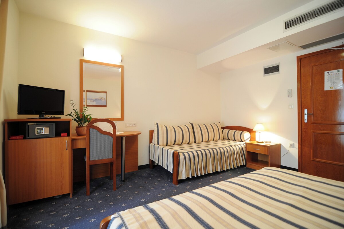 Una habitación de hotel con cama, escritorio con televisor, silla, mesita de noche con lámpara y puerta.