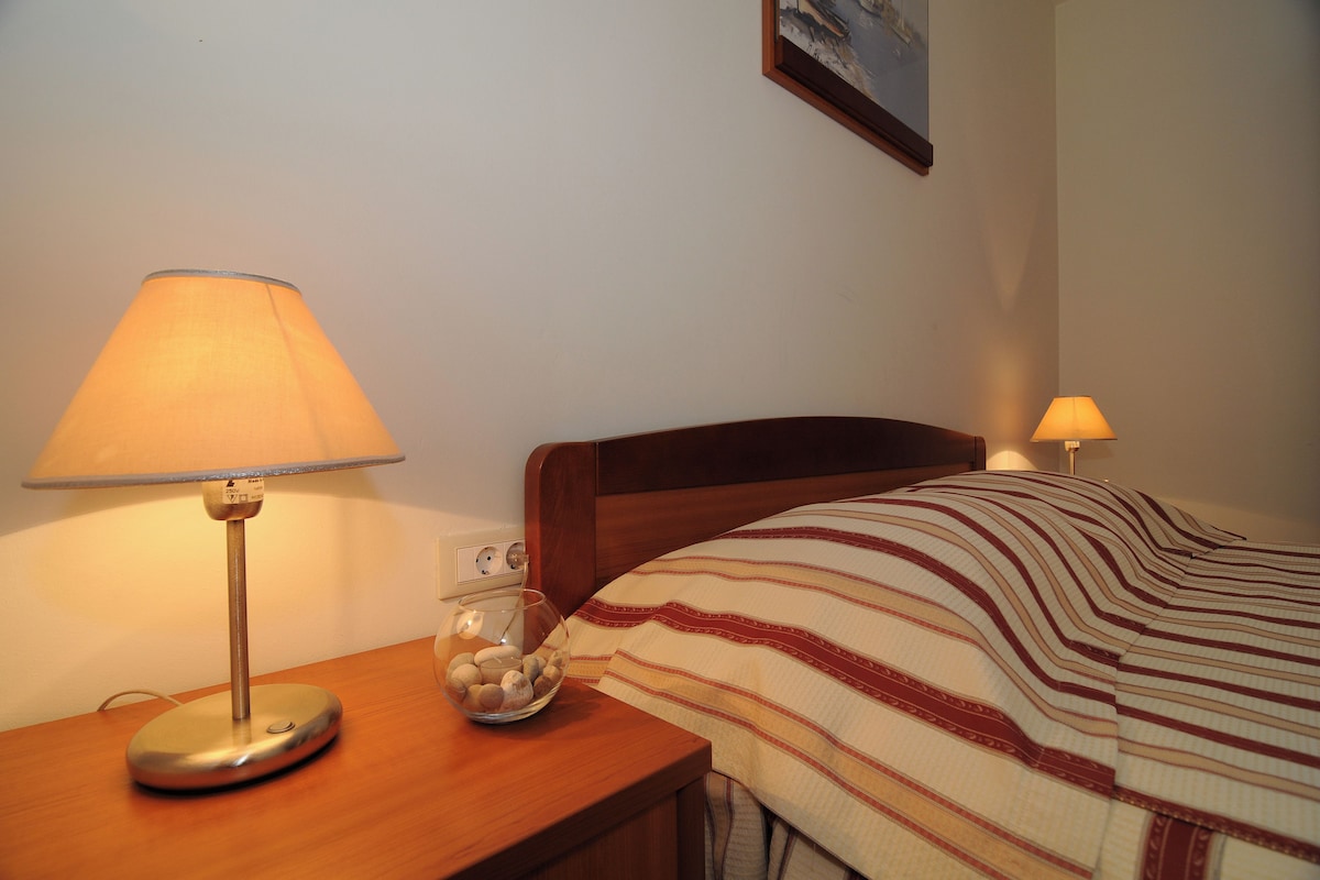Ein ordentlich bezogenes Bett mit einer gestreiften Decke, einer Nachttischlampe und einer Schale mit Steinen auf einem Holznachttisch.