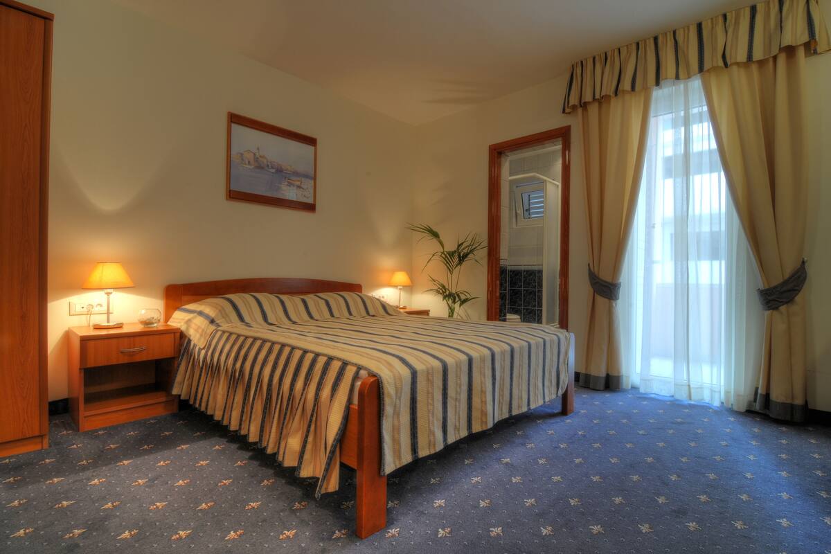 A hotel room with a bed, two bedside lamps, a nightstand, a mirror, a window with curtains, and a wall-mounted picture.