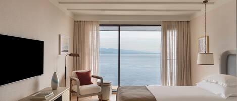 A hotel room with a large bed, a TV, a chair, and a view of the sea.