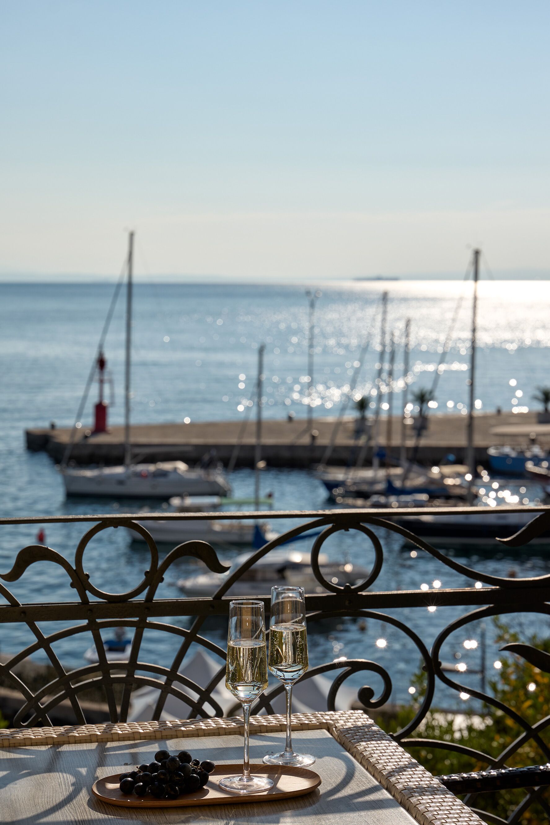 A table with two glasses of white wine and a plate of black olives, overlooking a marina with boats and a pier.