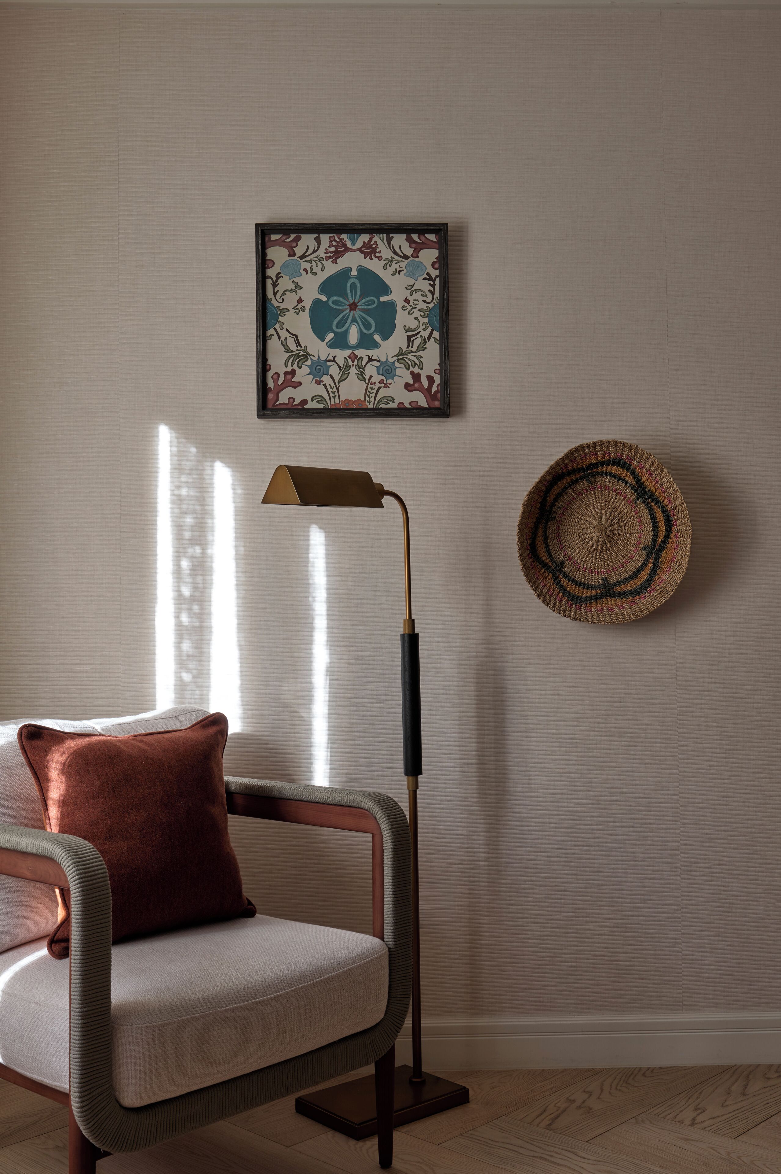 Una sala moderna con un sillón acolchado, una lámpara de pie, una cesta tejida y una obra de arte enmarcada en la pared.