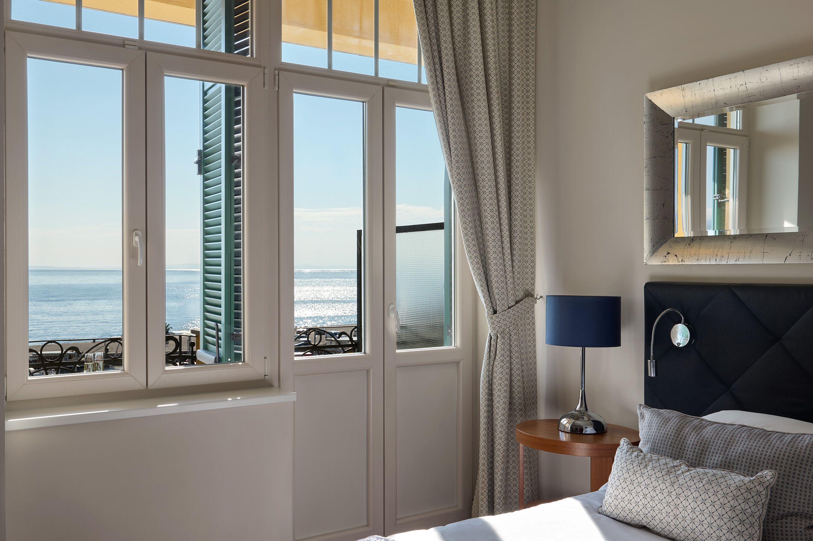 Habitación de hotel con cama, mesita de noche con lámpara, espejo y vistas al mar a través de ventanas abiertas.