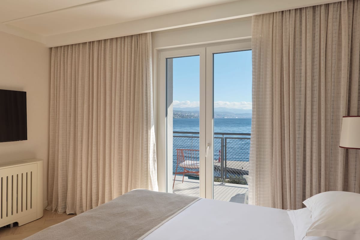 A hotel room with a bed, a TV, a view of the sea, and a balcony with a chair.