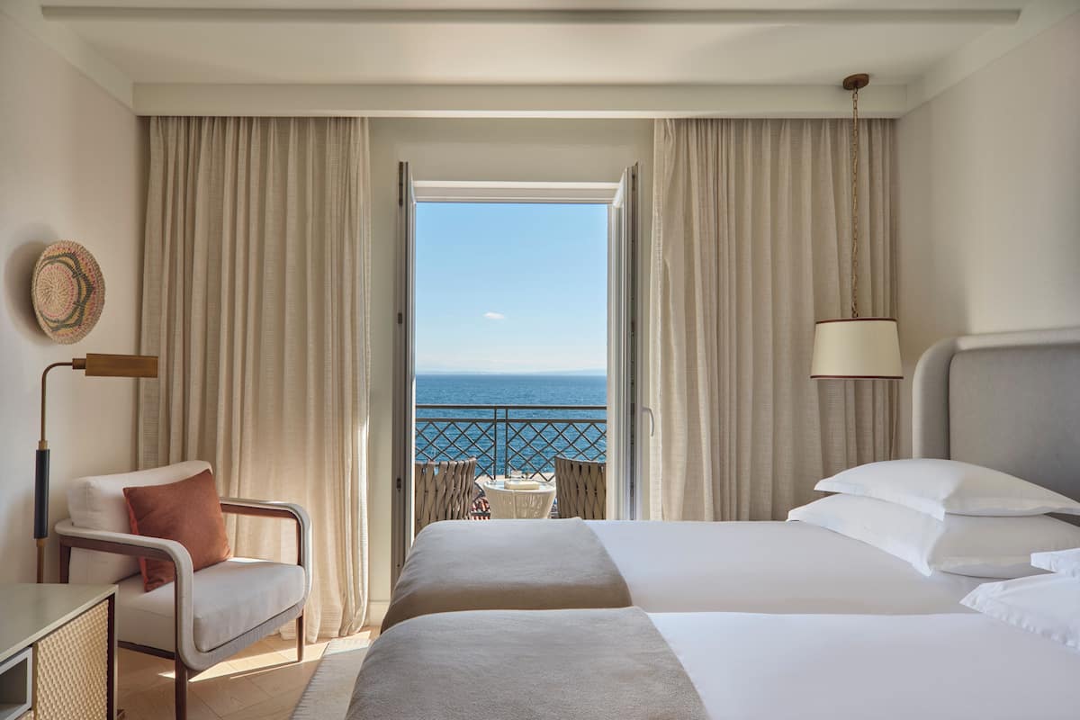 A hotel room with a bed, a chair, a small table, and a view of the sea through an open door.