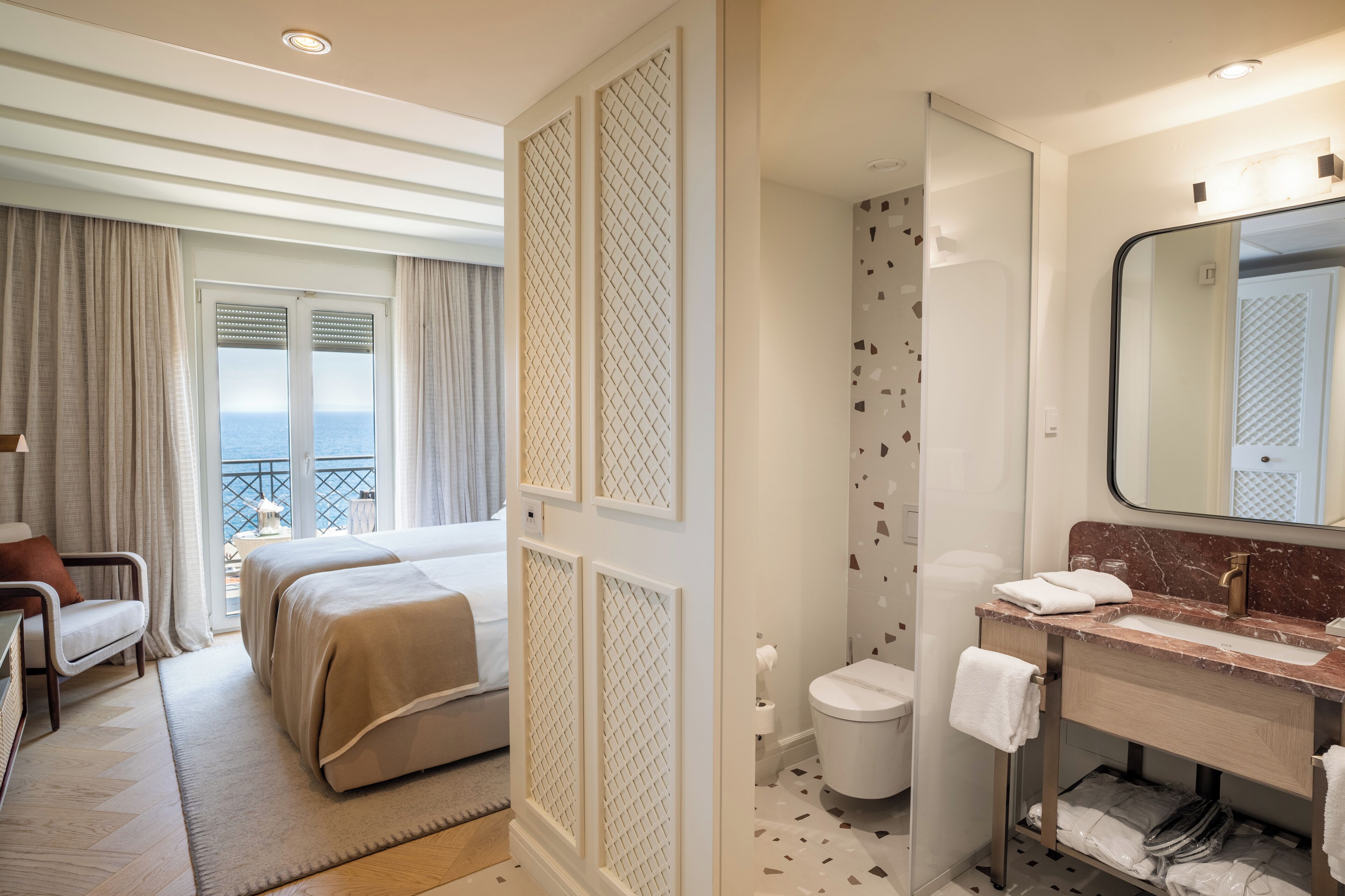 A hotel room with a bed, a desk, a chair, a bathroom with a sink and mirror, and a view of the sea through the window.