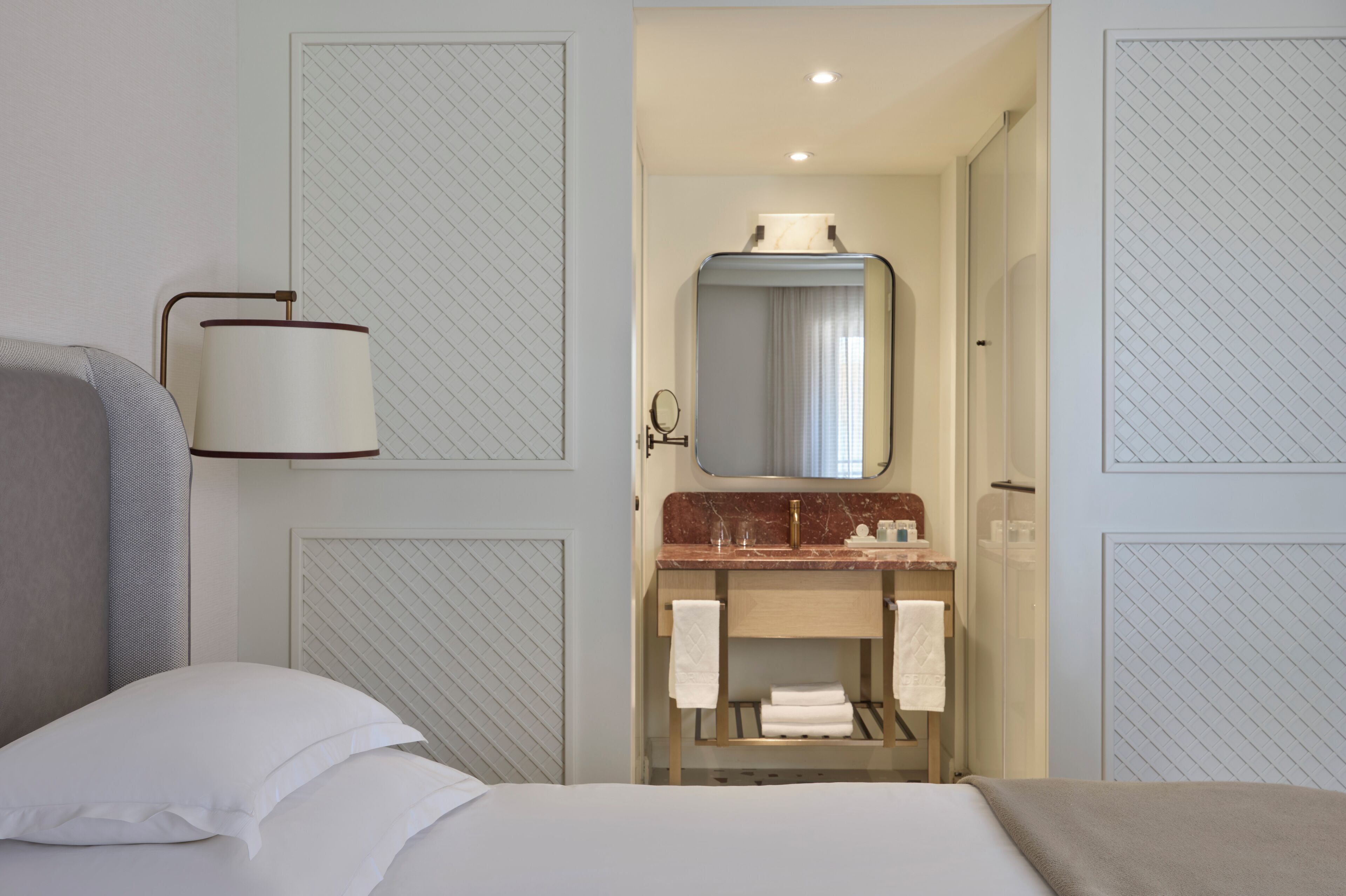 A hotel room with a bed, a bedside lamp, a bathroom with a sink, mirror, and towel rack.