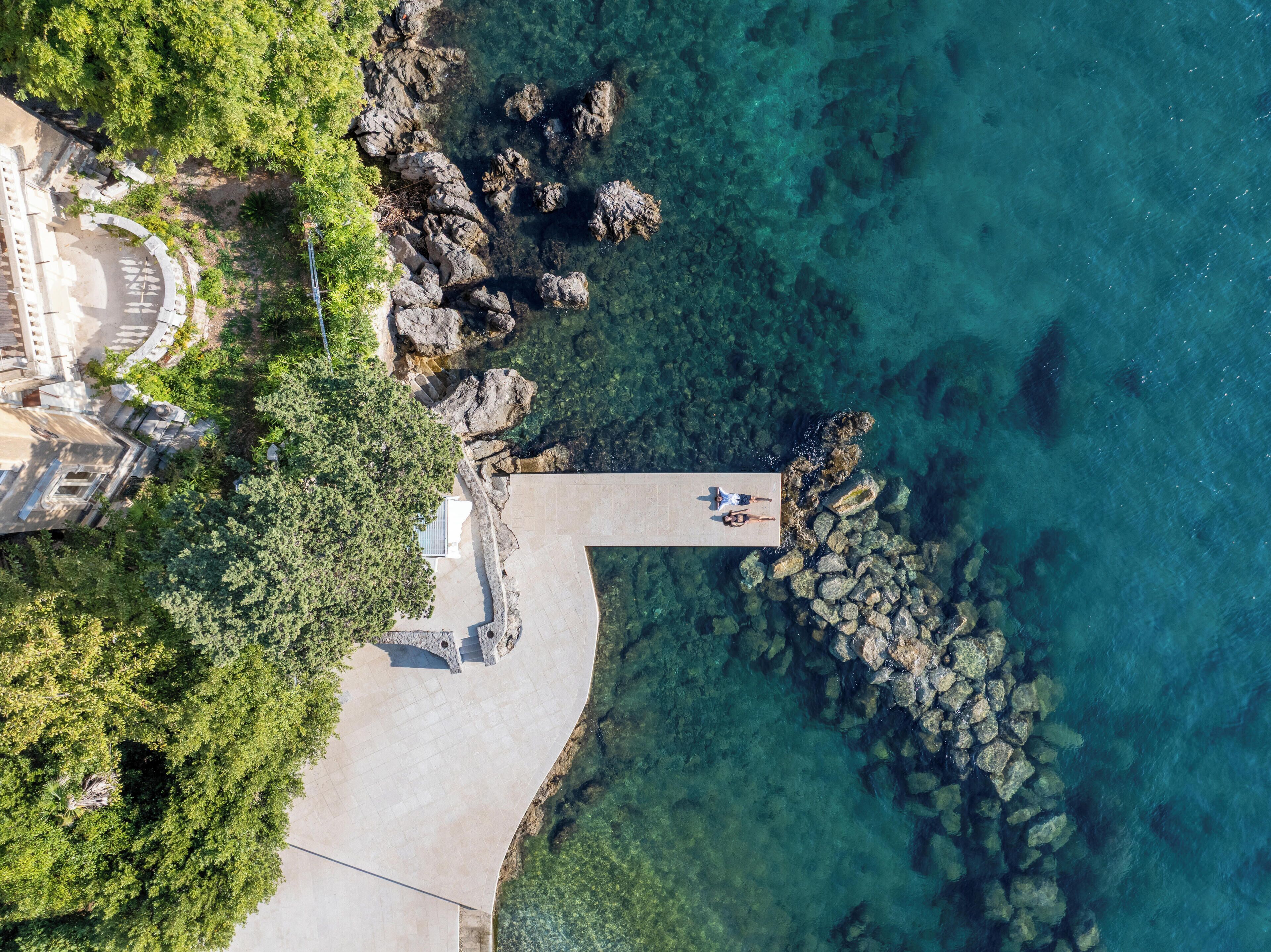 Vista aérea de una zona costera con un muelle que se adentra en aguas azules cristalinas, rodeada de formaciones rocosas y abundante vegetación.