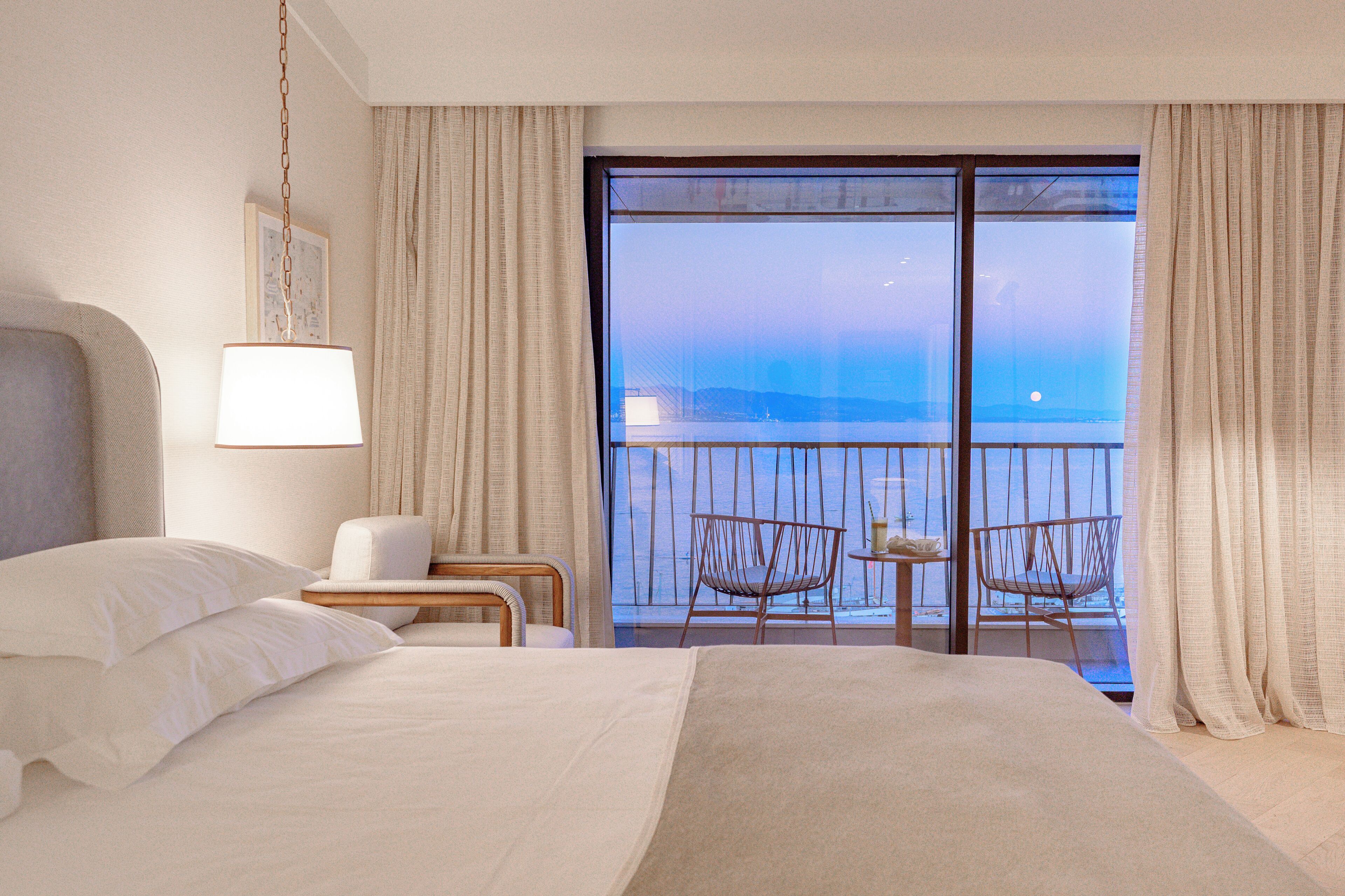 A hotel room with a large bed, a nightstand, a lamp, two chairs, and a balcony with a view.