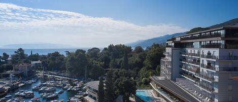 Un puerto deportivo con numerosas embarcaciones amarradas, un edificio grande con balcones con vistas al agua, y una vista despejada del mar y montañas a lo lejos.