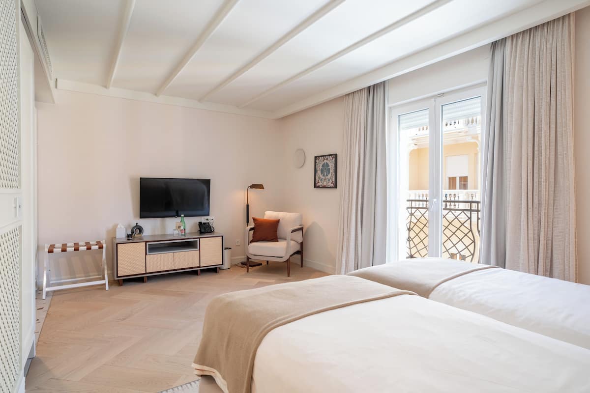 A hotel room with two beds, a TV, a desk with a lamp, and a view of the outside balcony.