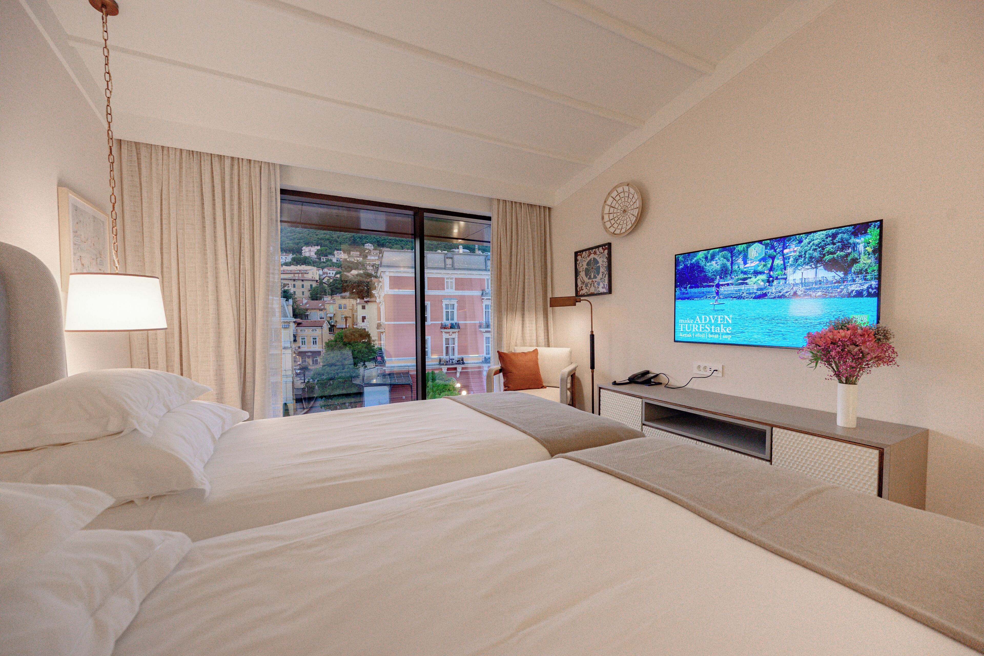 A hotel room with two beds, a TV, a desk, and a view of buildings through the window.