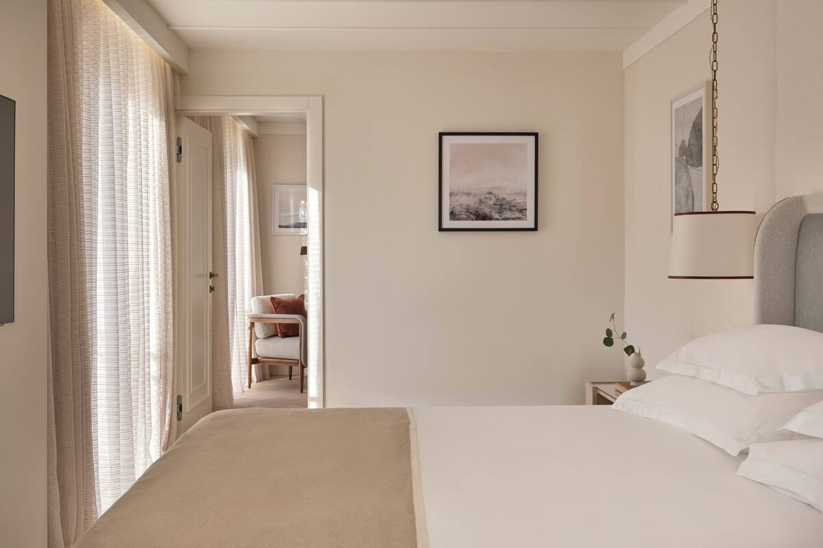 A hotel room with a bed, a nightstand, a framed picture on the wall, and a door leading to another room.