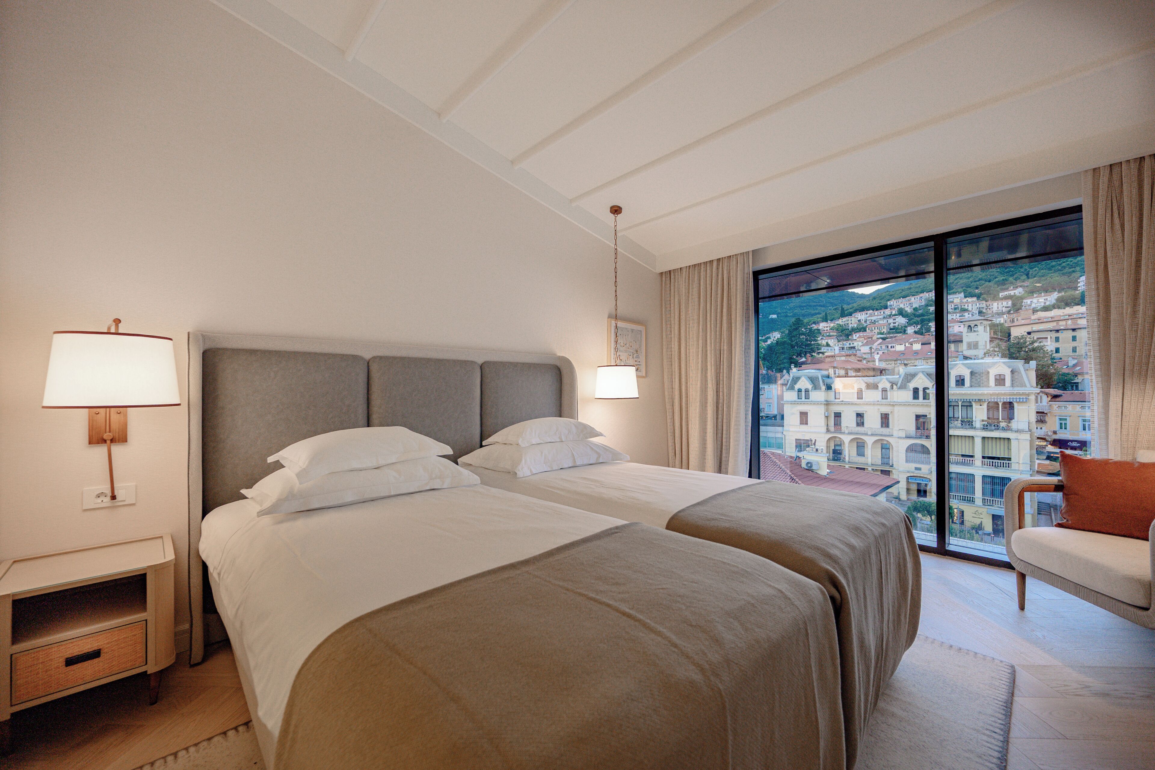 A hotel room with two beds, a nightstand, a lamp, and a view of a cityscape through large windows.