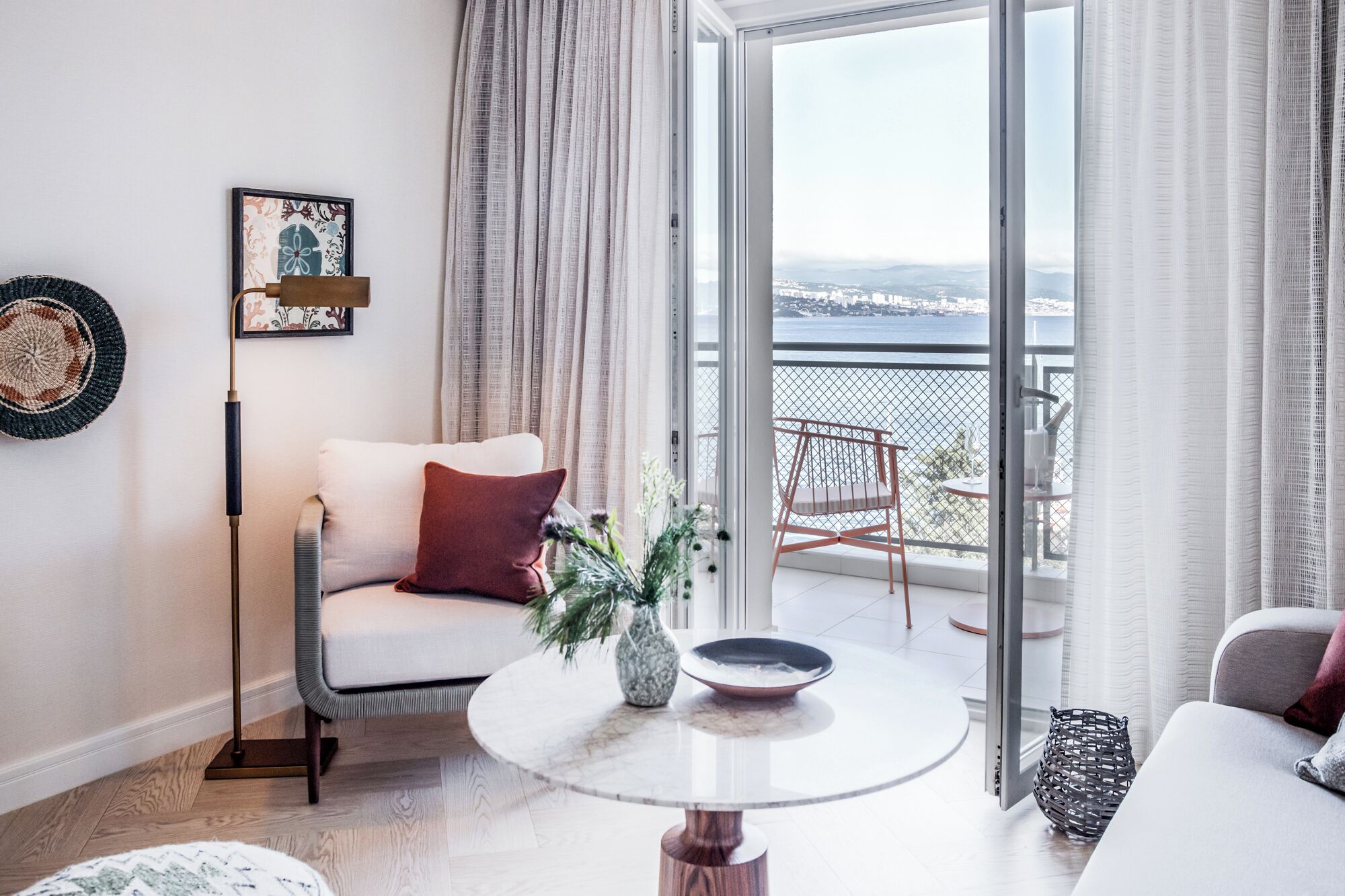 A modern living room with a sofa, a coffee table, a floor lamp, and a view of the sea through a balcony.
