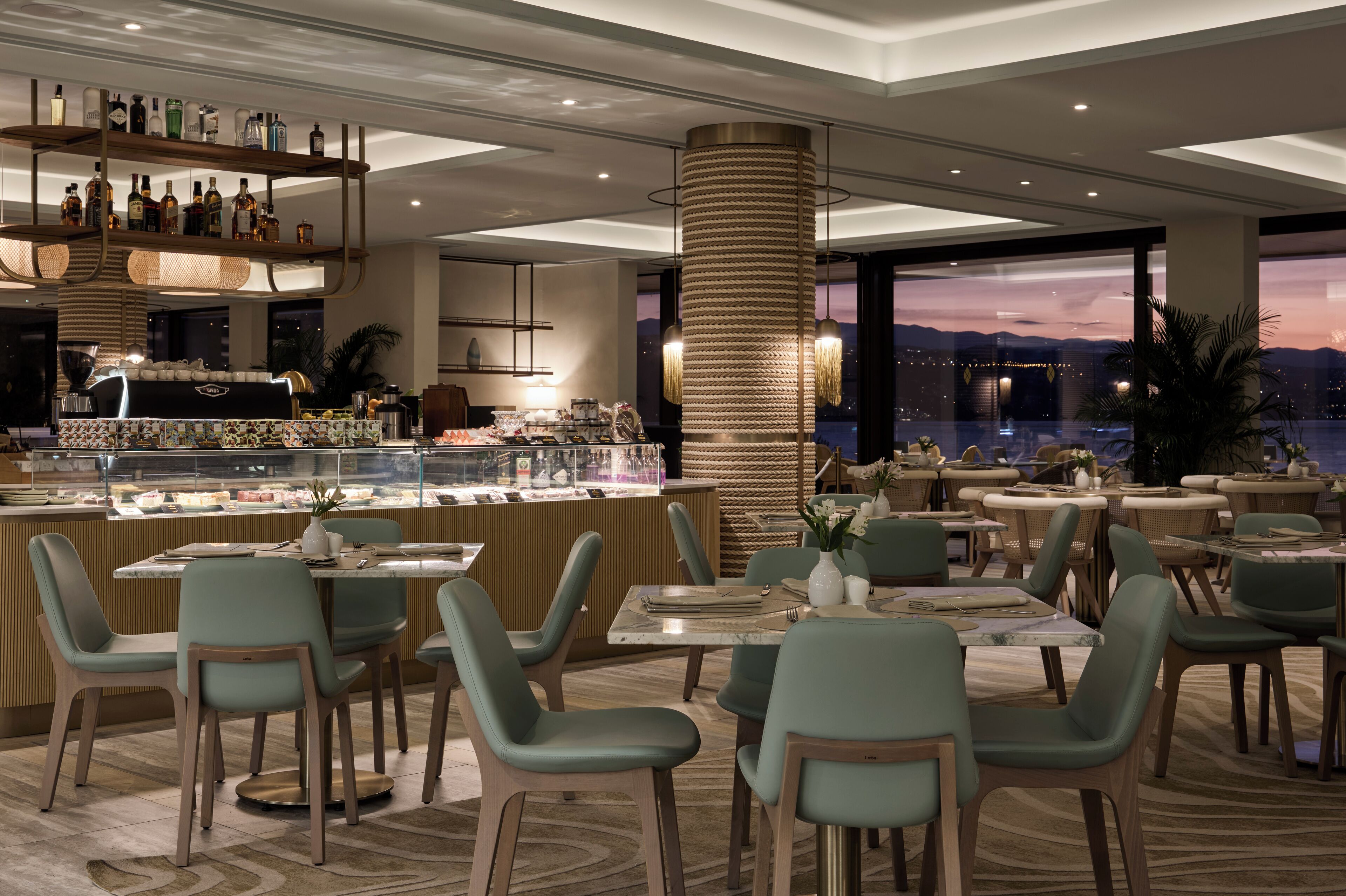 A modern restaurant with a bar area, multiple dining tables, and chairs, featuring a city view at dusk.