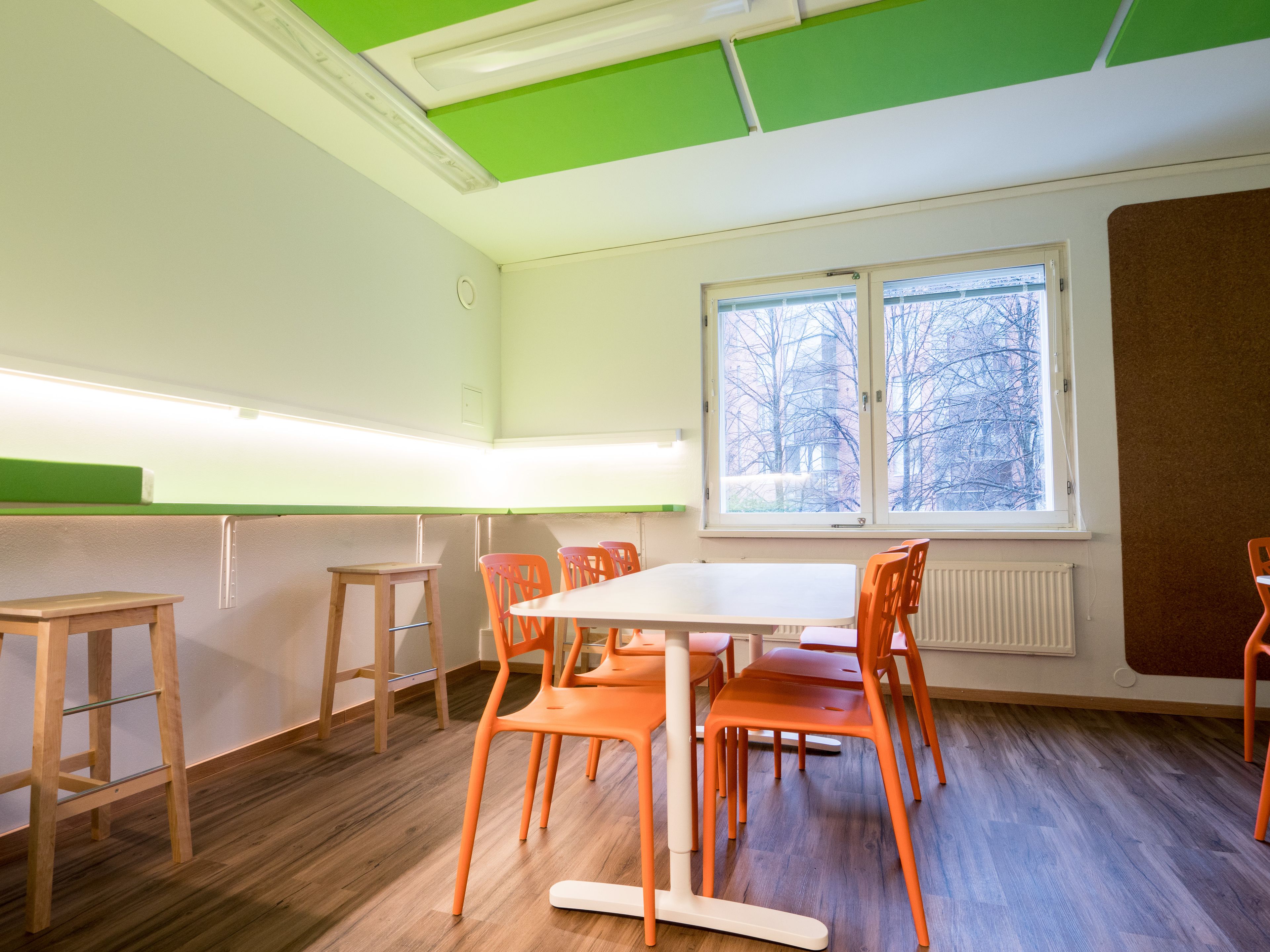 Een klaslokaal met een witte tafel, oranje stoelen en een raam met uitzicht op bomen.