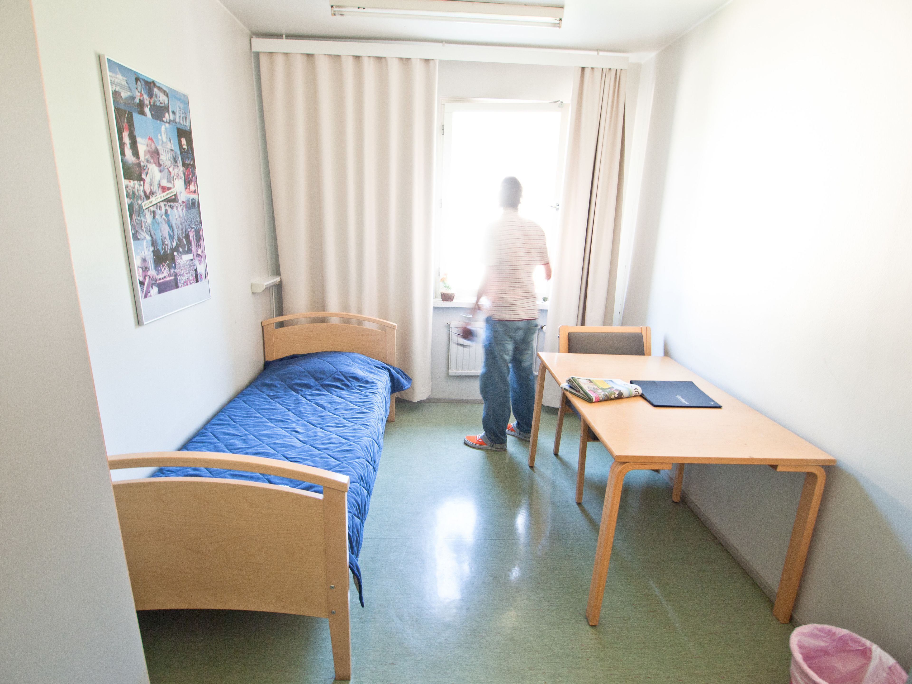 A small, single-bed room with a wooden headboard, a blue bedspread, a wooden desk, and a chair. There is a window with curtains, a radiator, and a poster on the wall.