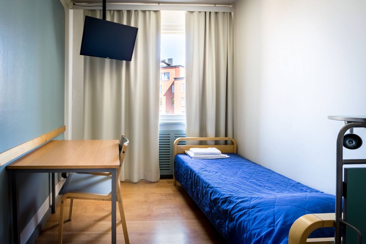 A small, tidy room with a wooden desk, a single bed with a blue bedspread, a television mounted on the wall, and a window with curtains.