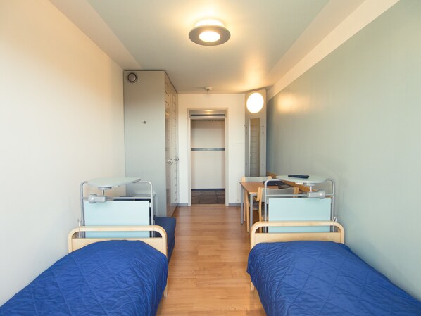 A room with two single beds, a small table, and a chair. There is a door leading to a bathroom.