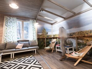 A cozy room with a wooden ceiling, a sofa, a chair, and a large window with a view of a lake and forest.