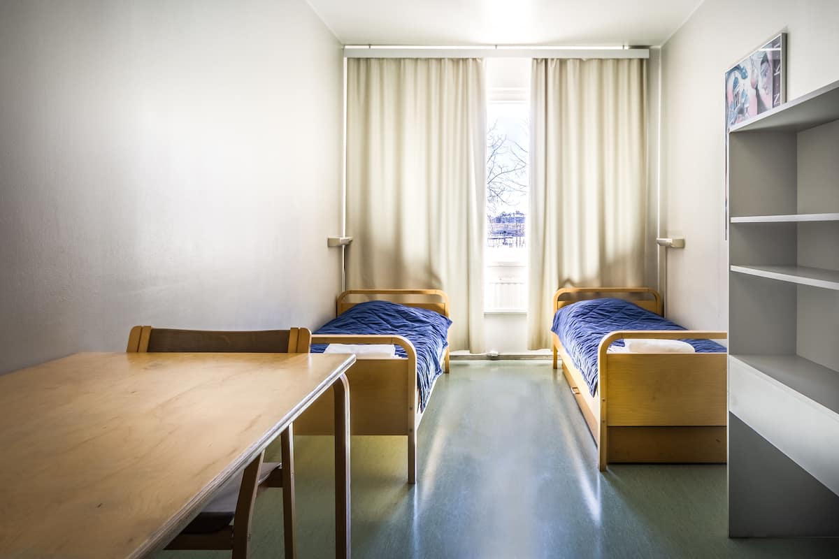 A small, clean room with two single beds, a wooden table, and a window with curtains.
