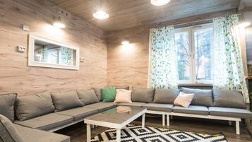 Una sala de estar acogedora con techo de madera, un ventanal con cortinas de hojas verdes, un sofá seccional gris con muchos cojines y una alfombra estampada.
