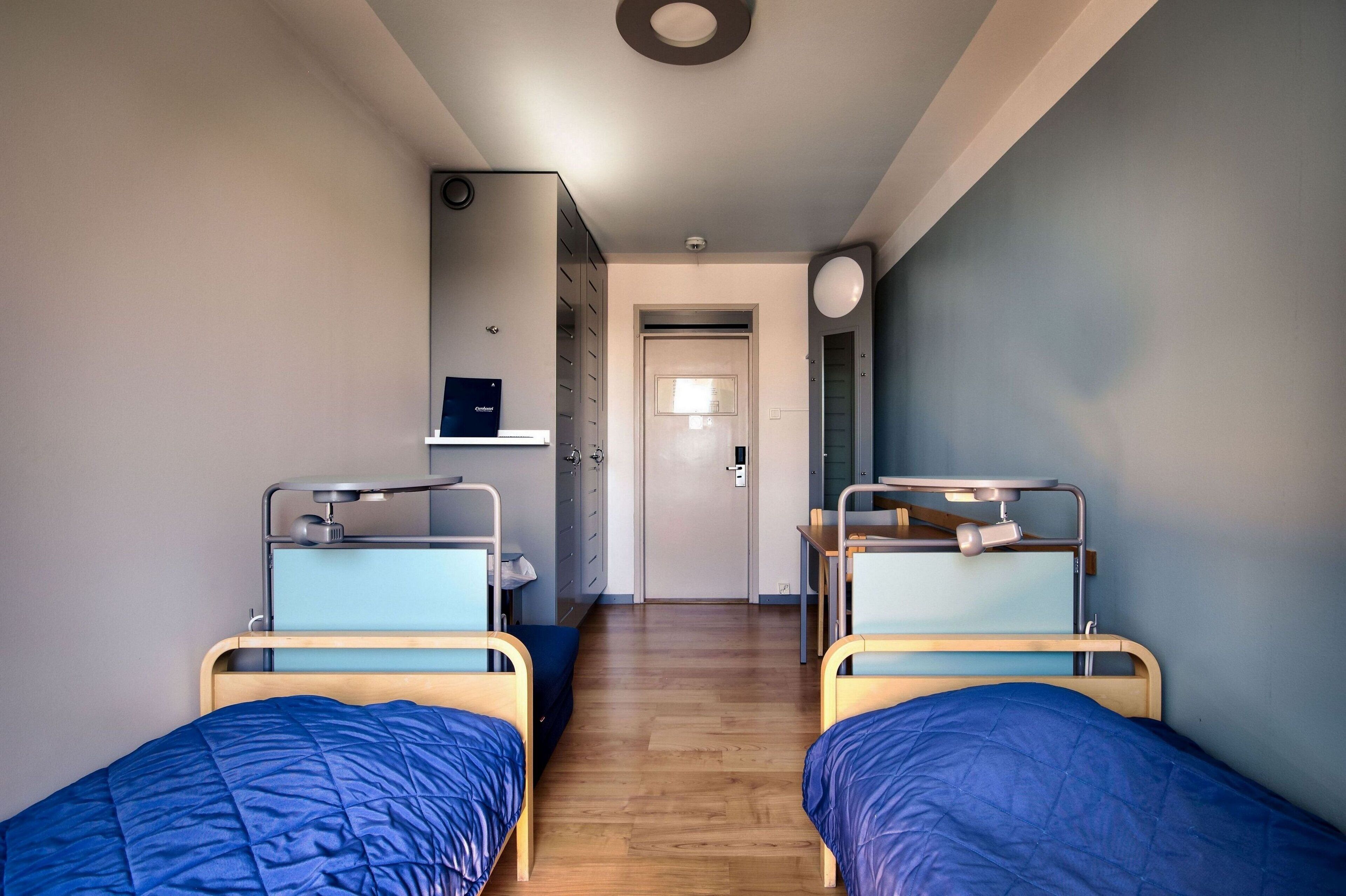 A narrow hallway with two single beds, a door at the end, and a small table with a lamp.