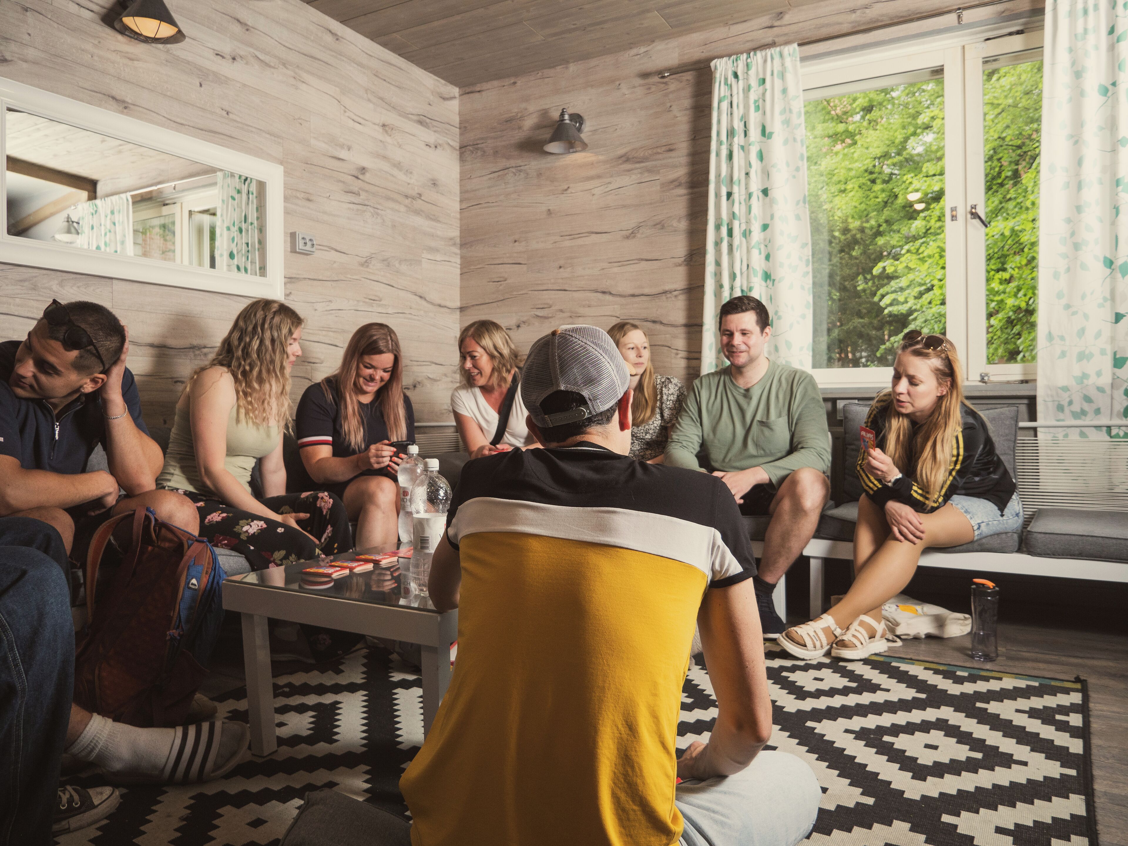 A group of people sitting on a couch and chairs in a room with wooden walls and a window with curtains.
