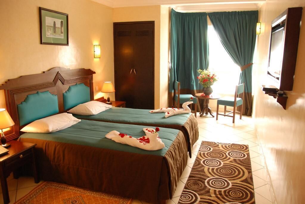 A hotel room with two beds, a desk, a chair, a small table with a vase, and a TV.