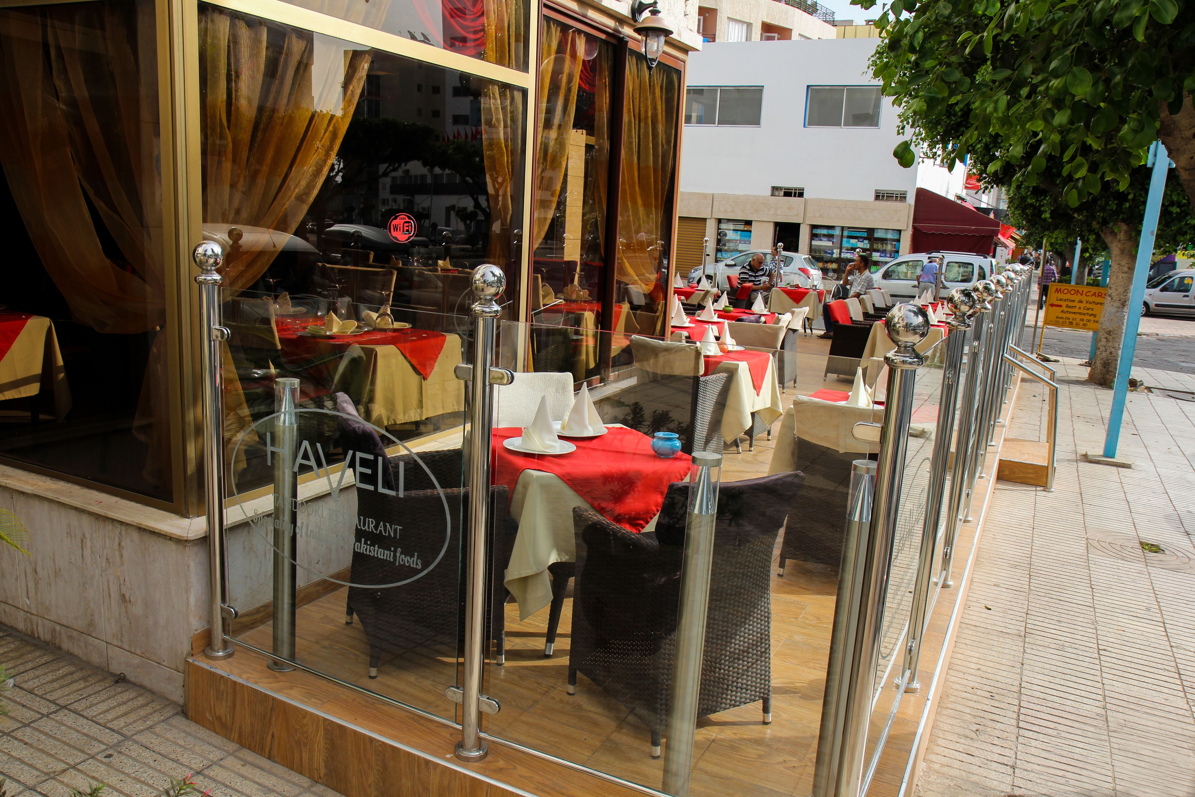 Un restaurante con terraza, barandillas de cristal y mantel rojo.