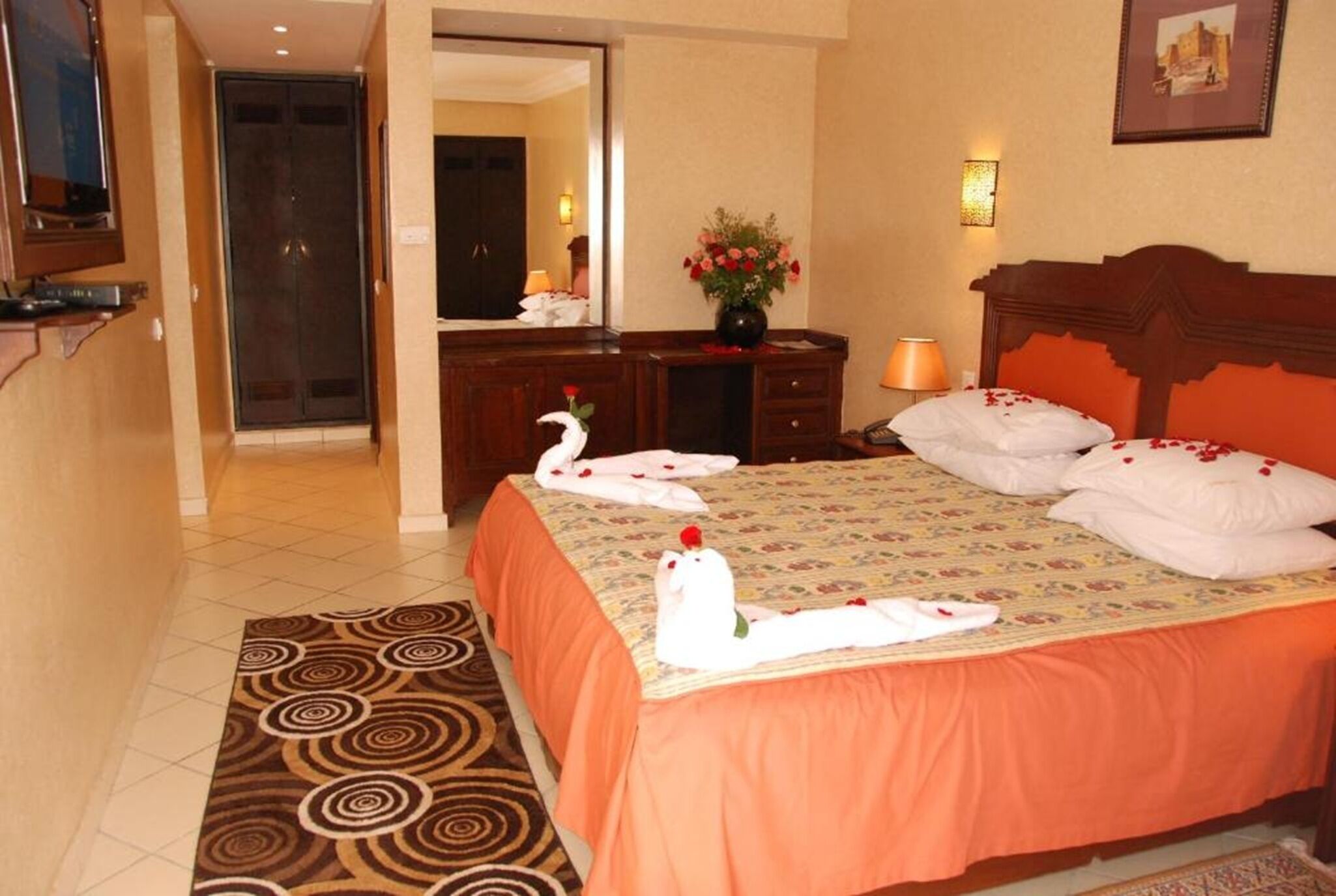 A hotel room with a bed, a dresser, a mirror, a vase with flowers, and a television.