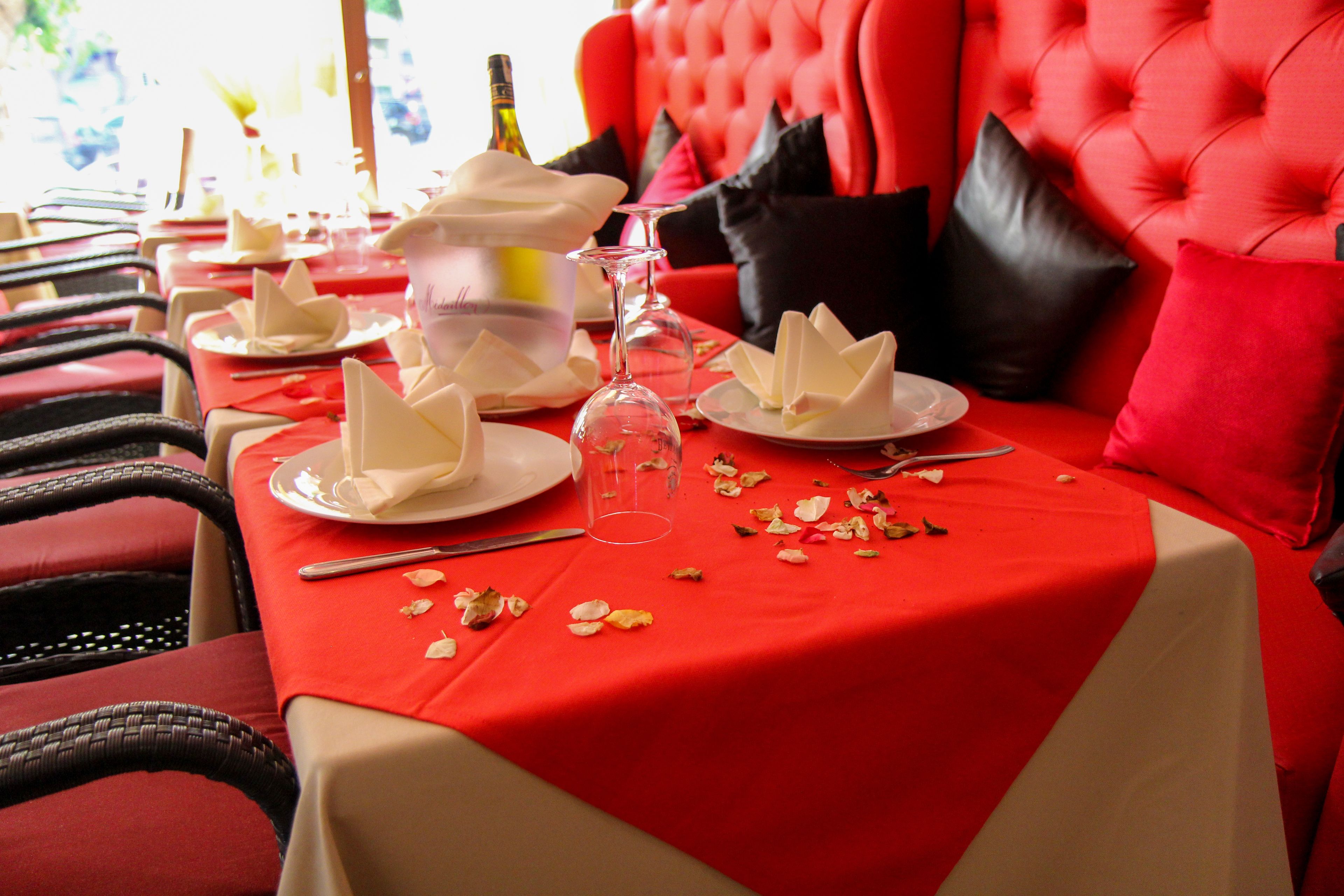 Una mesa de comedor dispuesta con servilletas blancas, una copa de vino y pétalos de rosa esparcidos.