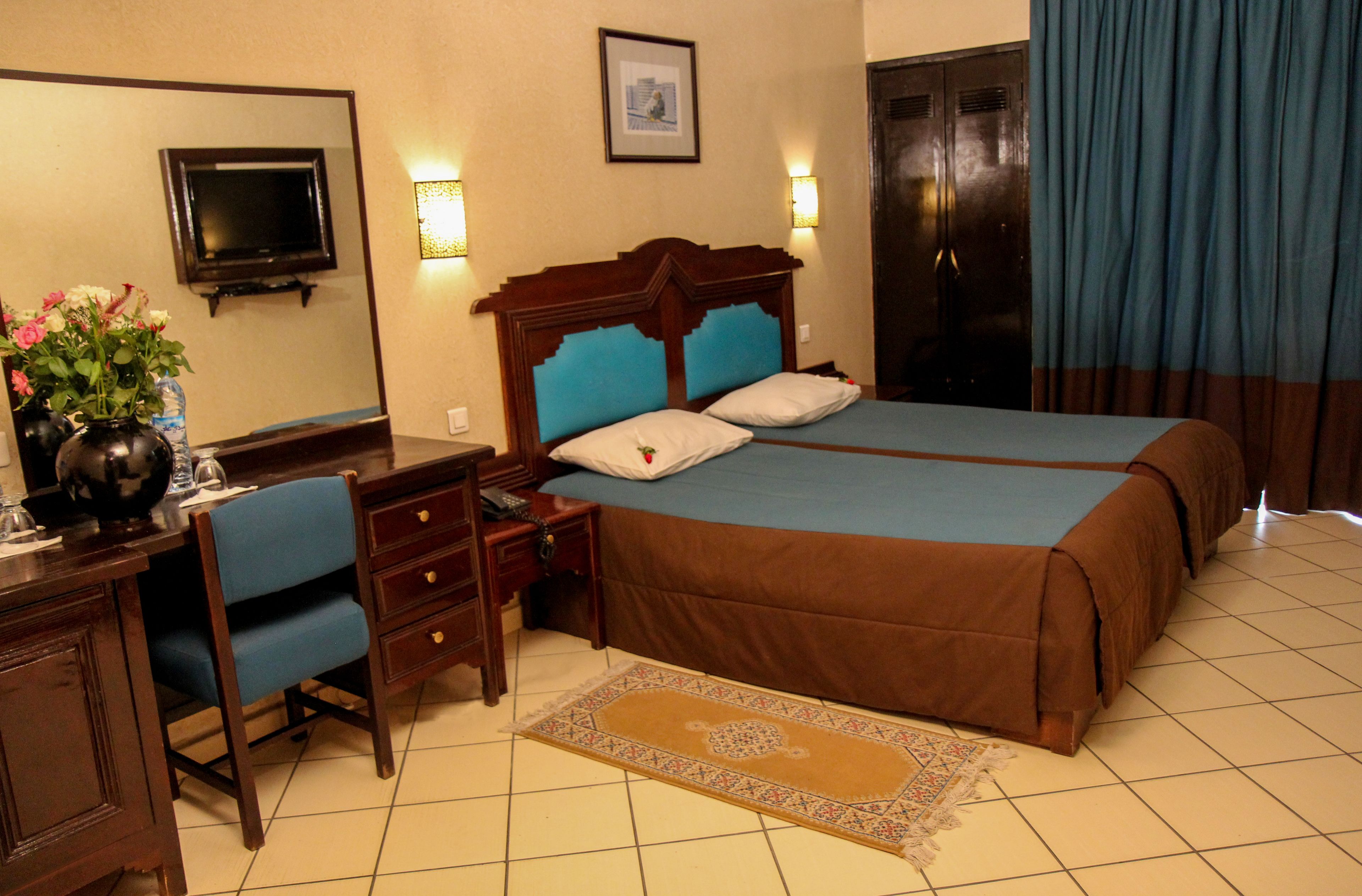 A hotel room with a bed, desk, chair, and a small table with a vase of flowers.