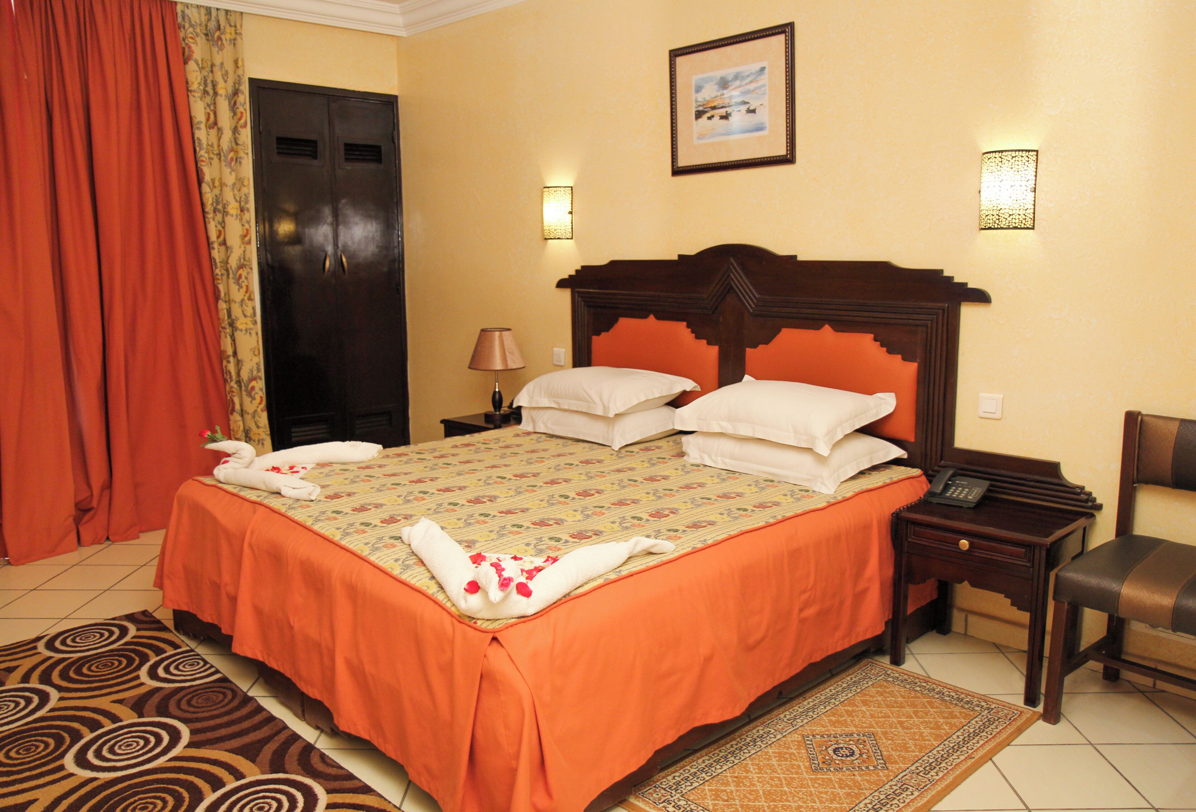 A hotel room with a large bed, a wooden headboard, a nightstand with a lamp, a chair, and a door with a curtain.