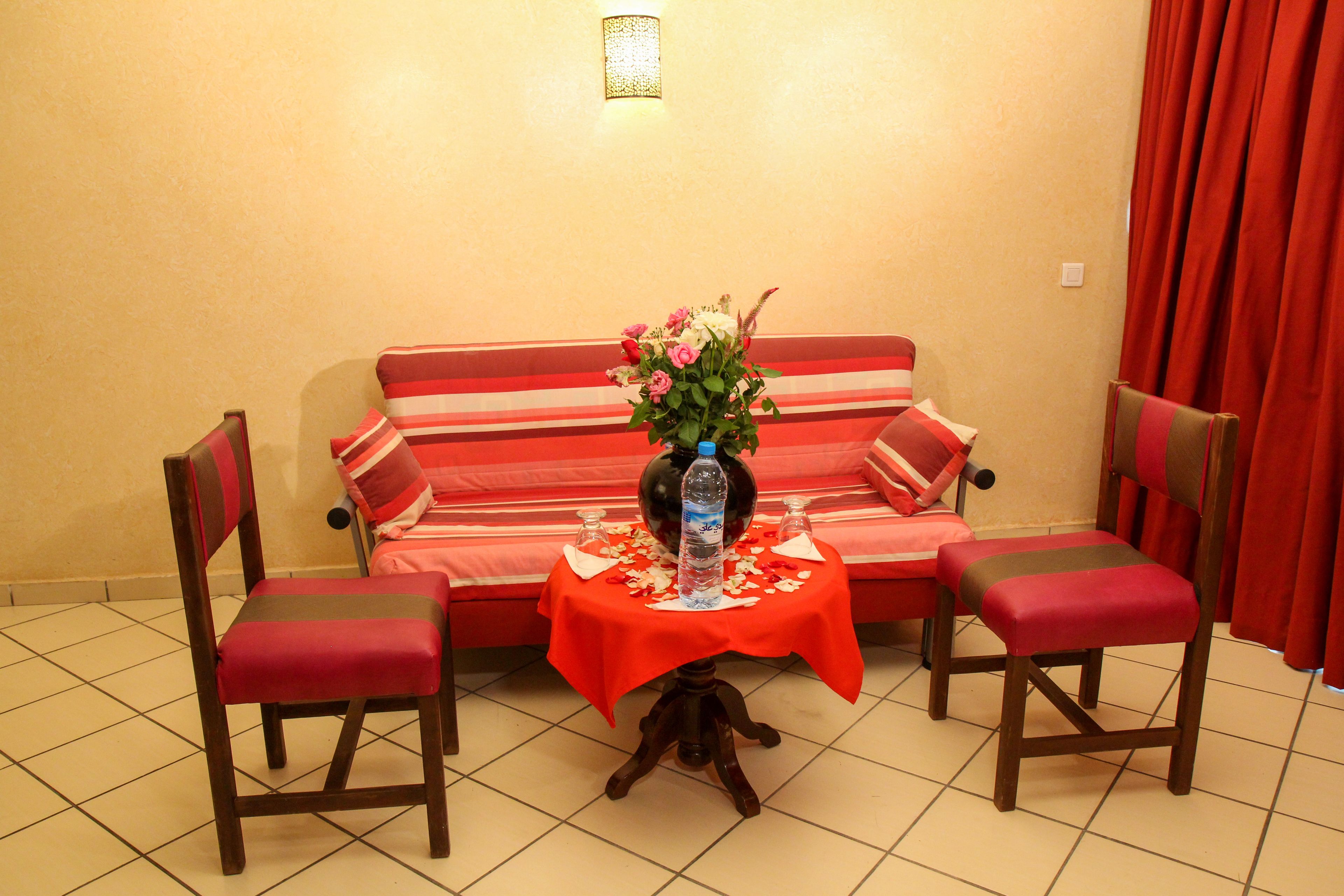 A room with a sofa, two chairs, a small table with a vase of flowers, and a bottle of water.