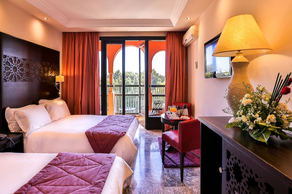 A hotel room with two beds, a desk with a lamp, a chair, and a view of the outdoors.