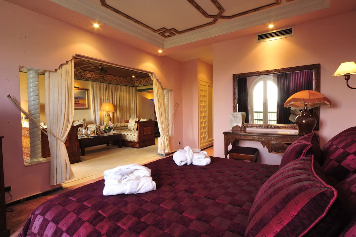 A hotel room with a large bed, a sitting area with a desk and chair, a mirror, and a view of the hallway.