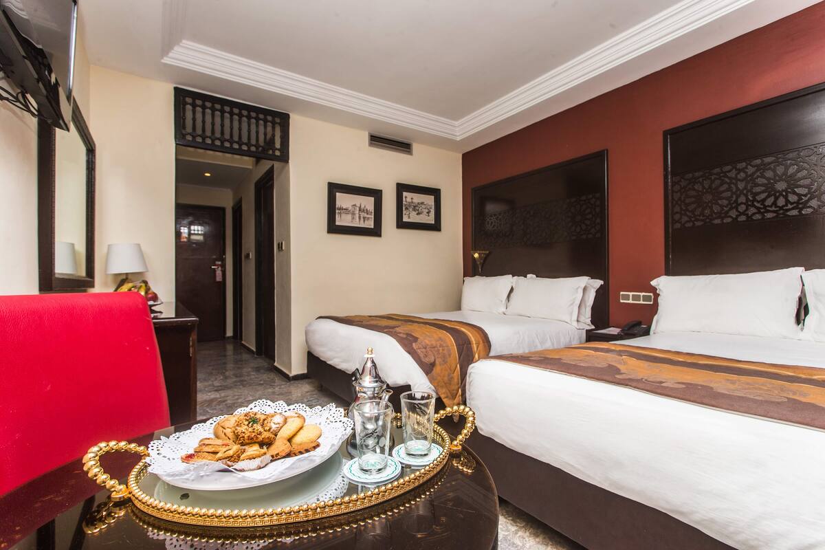 A hotel room with two beds, a dining table with a plate of pastries, and a red chair.