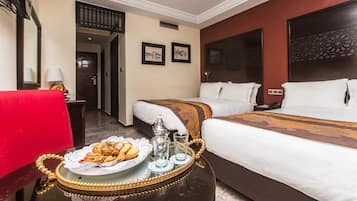 A hotel room with two beds, a dining table with a plate of pastries, and a red chair.