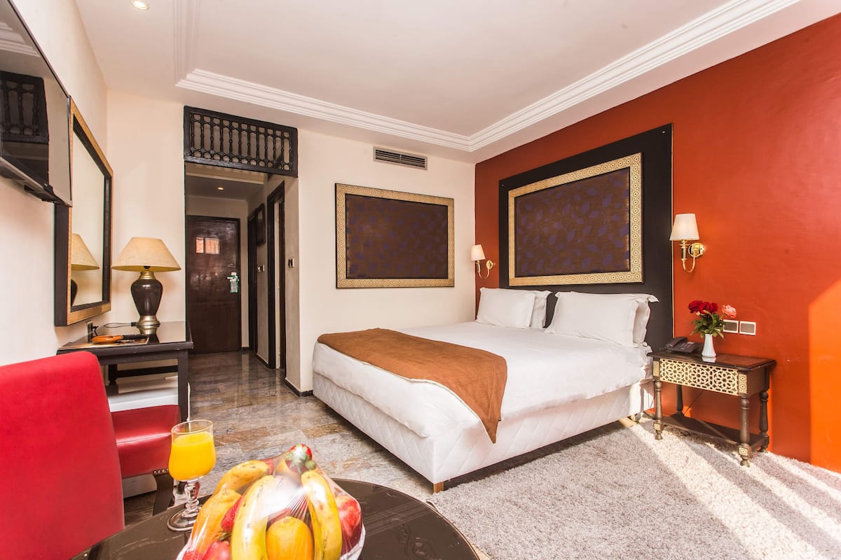 A hotel room with a bed, a desk with a lamp, a chair, a fruit bowl, and a nightstand with a vase.